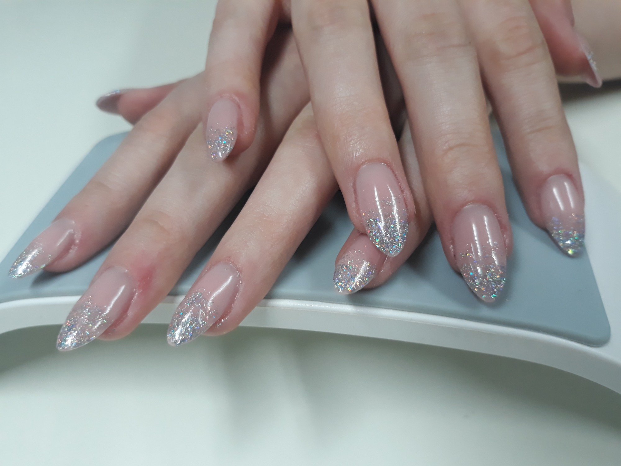 Dłonie kobiety z przedłużonymi, owalnymi paznokciami w kolorze nude z srebrnym brokatem na końcach, ułożone na szarym podłożu.