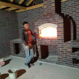 Exspress partner sp.z o.o. - Mężczyzna opiera się o ceglany piec zewnętrzny z palącym się ogniem wewnątrz, widoczne niedokończone elementy konstrukcyjne i wiadro z zaprawą.