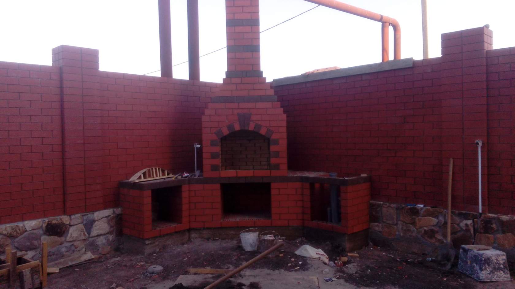Budowa murowanego grilla z czerwonej cegły z elementami z ciemniejszej cegły, widoczny etap prac z niezagospodarowanym terenem wokół, fragment muru ogrodzeniowego w tle.
