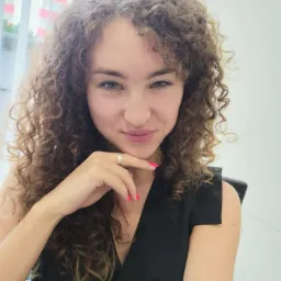 Portret uśmiechniętej kobiety z kręconymi włosami, opierającej podbródek na dłoni z różowym manicure, w czarnej bluzce na tle okna i białej ściany.