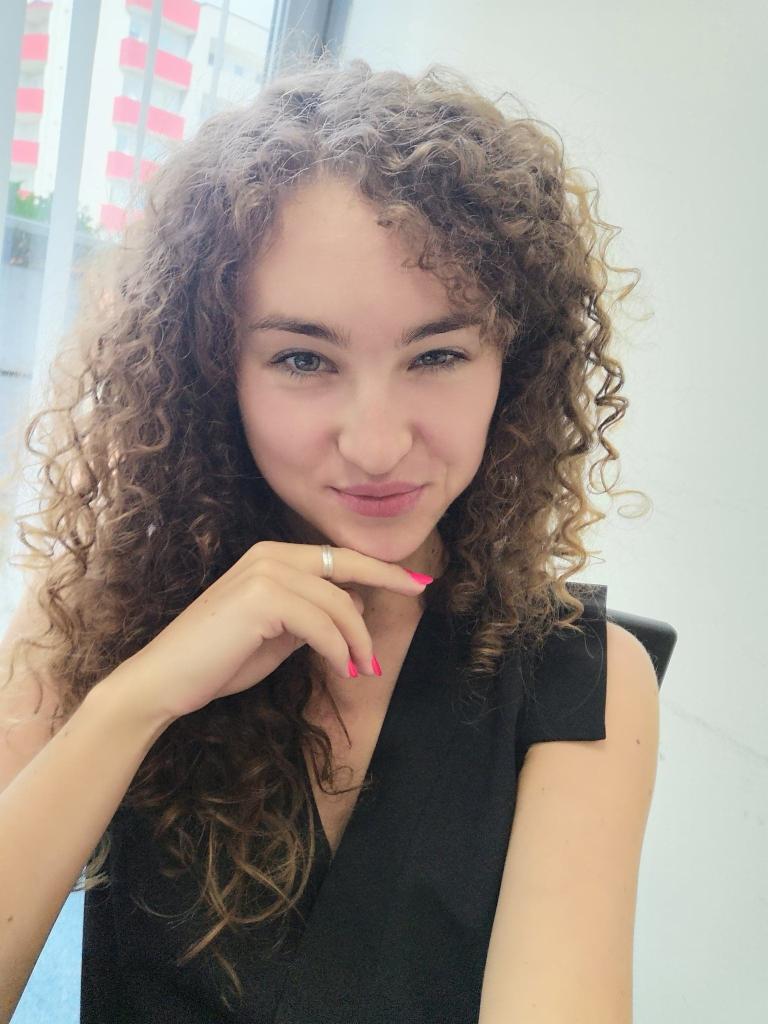 Portret uśmiechniętej kobiety z kręconymi włosami, opierającej podbródek na dłoni z różowym manicure, w czarnej bluzce na tle okna i białej ściany.