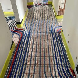 BUDOTOM SPÓŁKA Z OGRANICZONĄ ODPOWIEDZIALNOŚCIĄ - Ułożone rury ogrzewania podłogowego w korytarzu, zabezpieczone żółtą taśmą przy ścianach. Instalacja na folii z czerwoną siatką. Widok z góry, przed wylaniem posadzki.