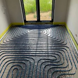 Niebieskie rury ogrzewania podłogowego ułożone równolegle na folii izolacyjnej w jasnym pokoju z oknem wychodzącym na zieleń. Żółta taśma wzdłuż ścian.