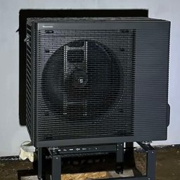 BUDOTOM SPÓŁKA Z OGRANICZONĄ ODPOWIEDZIALNOŚCIĄ - Czarna pompa ciepła Panasonic Aquarea na metalowym stelażu, zainstalowana przy białej ścianie budynku. Widoczna instalacja i ziemia wokół.