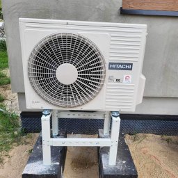 BUDOTOM SPÓŁKA Z OGRANICZONĄ ODPOWIEDZIALNOŚCIĄ - Zamontowana jednostka zewnętrzna klimatyzacji Hitachi na podwyższeniu z metalowych profili, na tle ściany budynku z oknem. Widoczne podłoże piaszczyste.