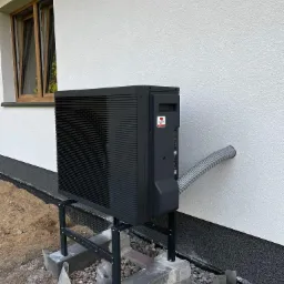 Czarna pompa ciepła Panasonic Aquarea zamontowana na zewnątrz budynku z białą elewacją. Widoczny szary przewód wchodzący w ścianę i okno z drewnianą ramą.