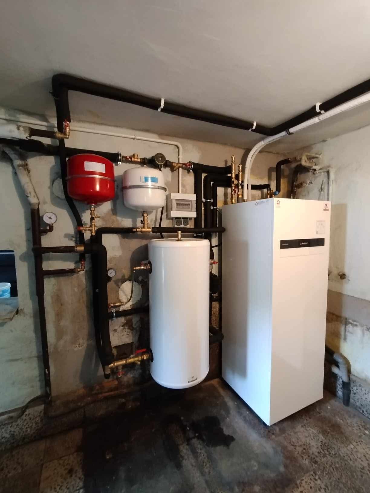 Biała pompa ciepła Viessmann z zbiornikiem i instalacją hydrauliczną w piwnicy. Widoczne rury w izolacji, manometry i zawory.