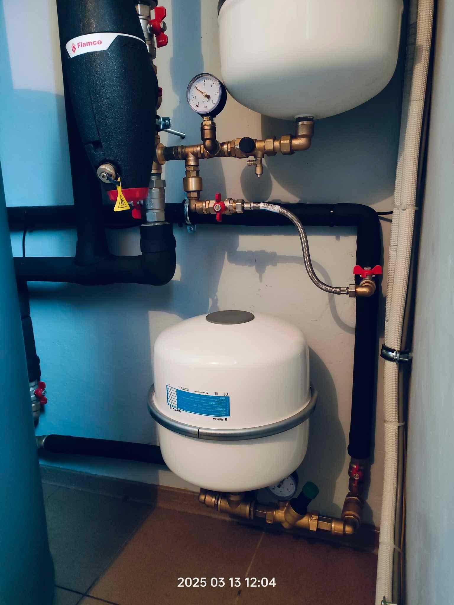 Zamknięty system grzewczy Flamco z czarnymi rurami, manometrem i białymi zbiornikami wyrównawczymi w pomieszczeniu technicznym. Widoczne mosiężne złączki i zawory.