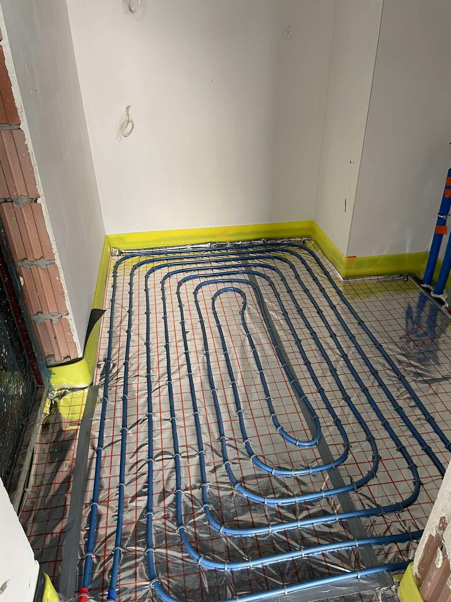 Niebieskie rury ogrzewania podłogowego ułożone na folii z siatką w pomieszczeniu przed wylaniem posadzki, widoczne elementy mocujące i żółta taśma brzegowa.