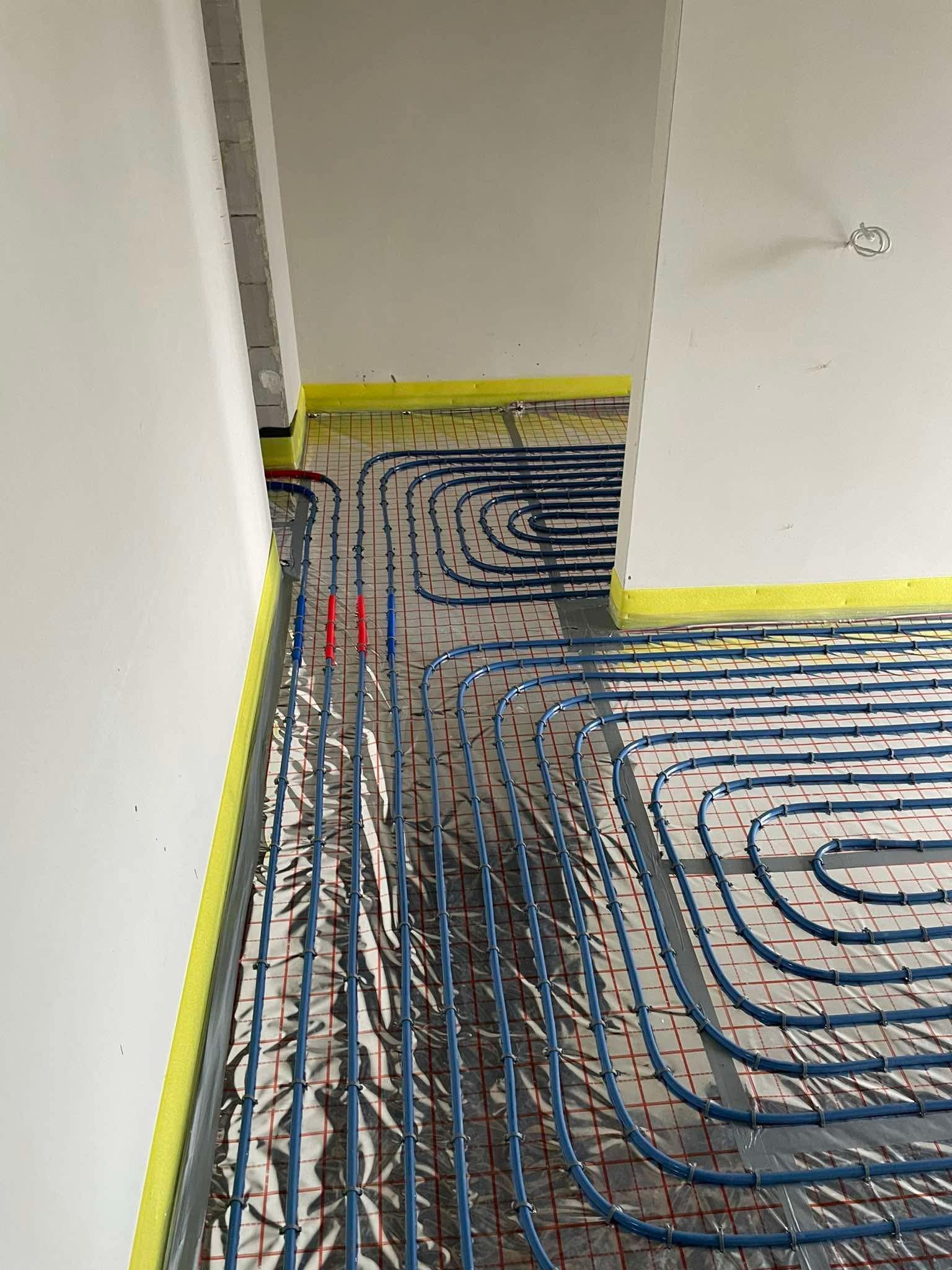 Niebieskie rurki ogrzewania podłogowego ułożone na folii z czerwoną siatką, zabezpieczone żółtą taśmą przy ścianach, w trakcie montażu w jasnym wnętrzu.