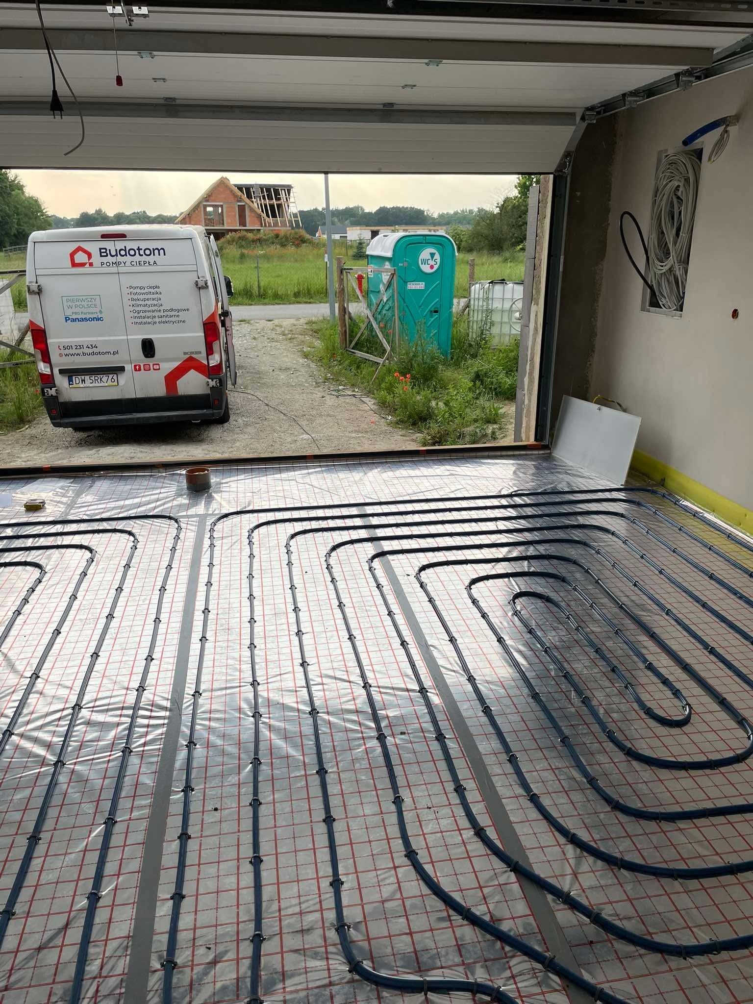 Rury ogrzewania podłogowego ułożone na folii z siatką, widok z garażu na budowę domu i auto firmy Budotom. Instalacja na podkładzie, etap przed wylewką.