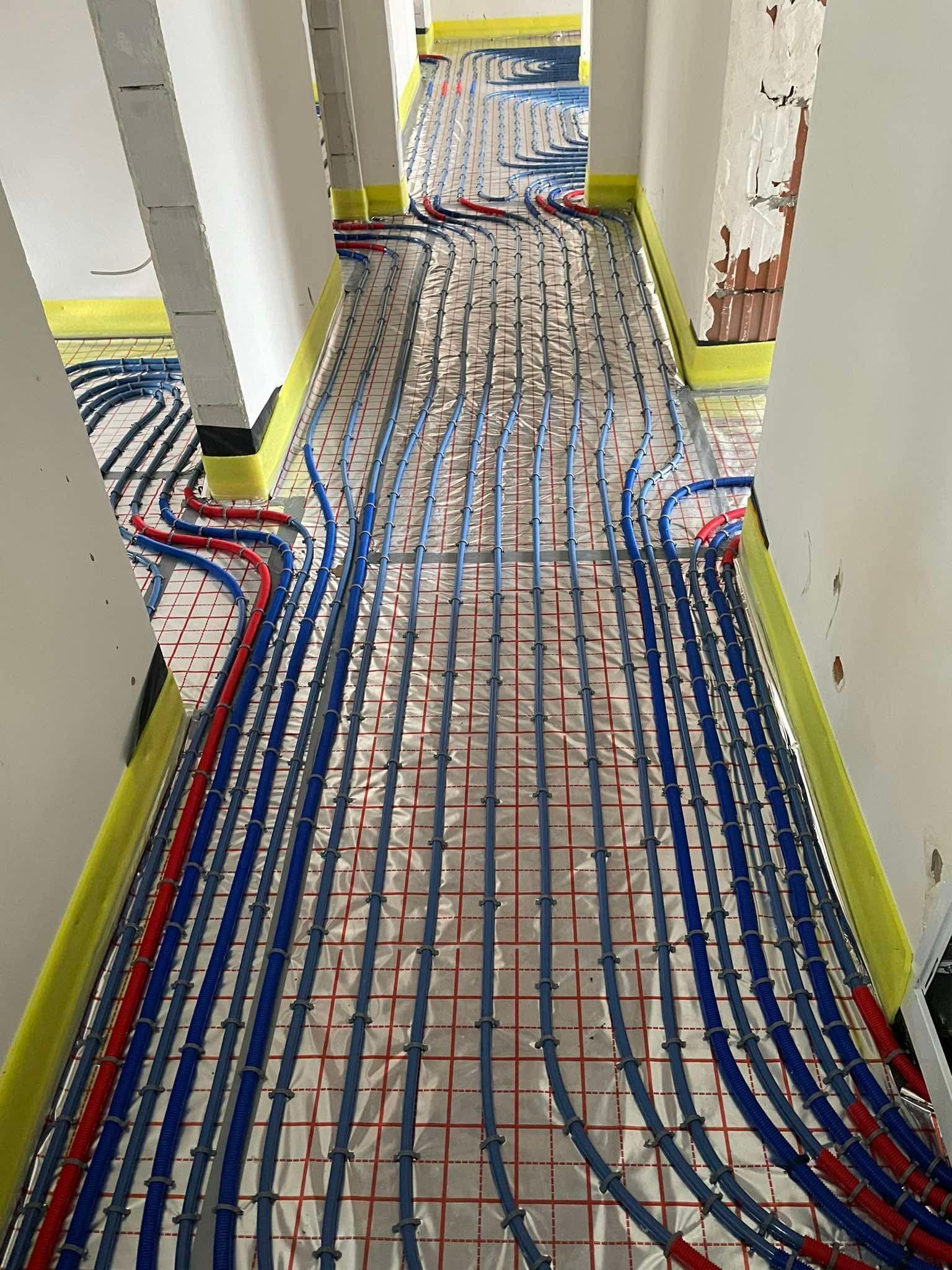Ułożone rury ogrzewania podłogowego w korytarzu, zabezpieczone żółtą taśmą przy ścianach. Instalacja na folii z czerwoną siatką. Widok z góry, przed wylaniem posadzki.