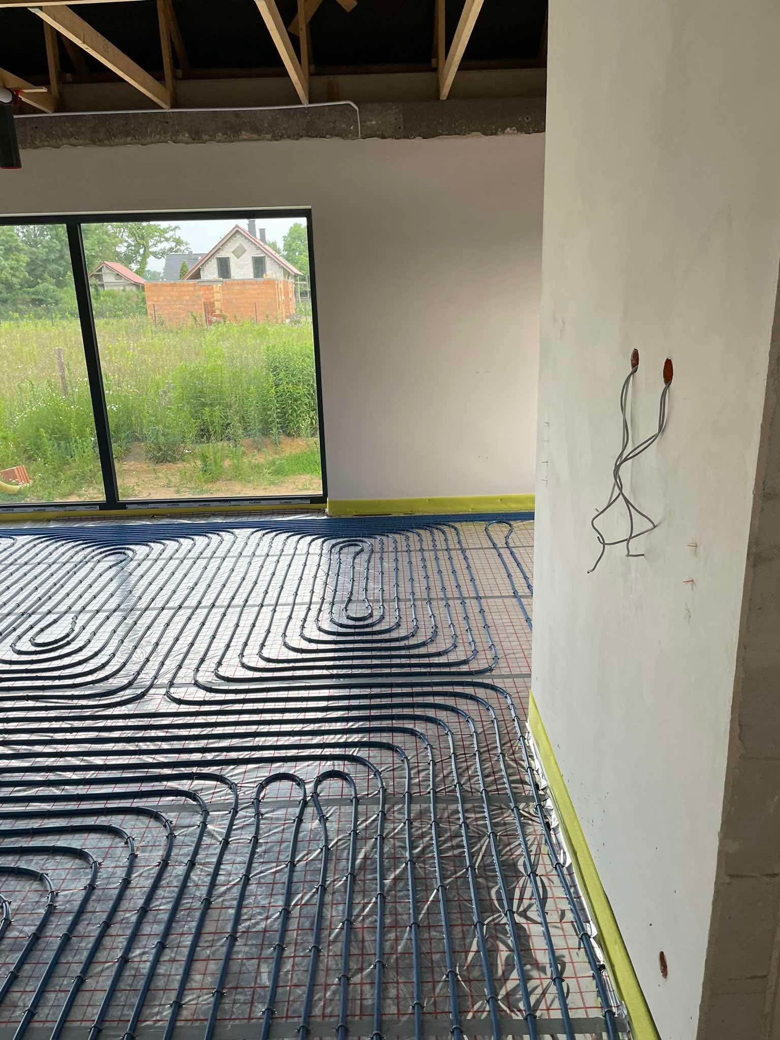 Niebieskie rury ogrzewania podłogowego rozłożone na folii z siatką w surowym wnętrzu domu z widokiem na ogród przez okno. Widoczne przewody elektryczne.