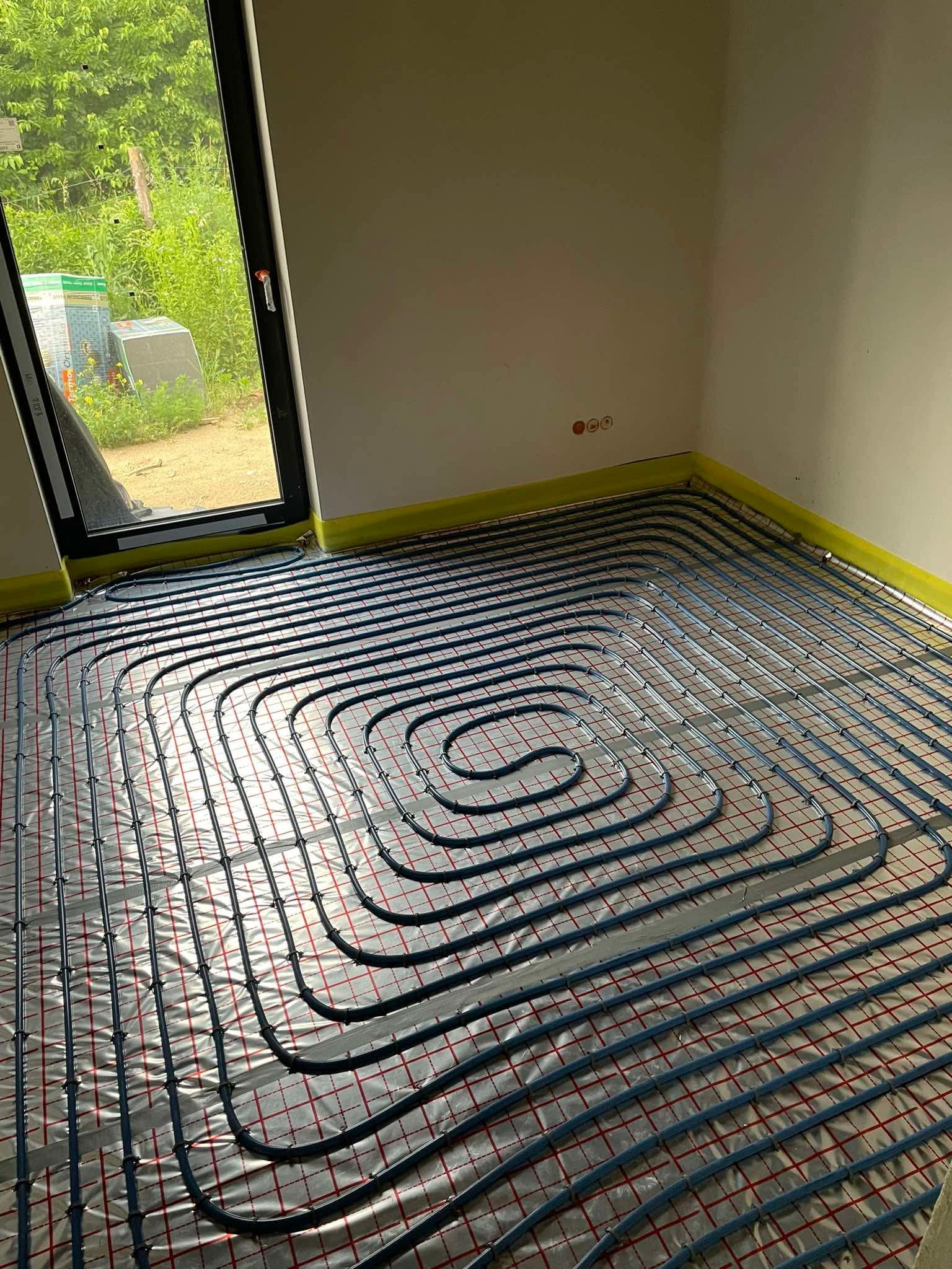 Niebieskie rury ogrzewania podłogowego ułożone spiralnie na srebrnej folii z czerwoną siatką w jasnym pomieszczeniu. Widoczne okno i fragment ściany.