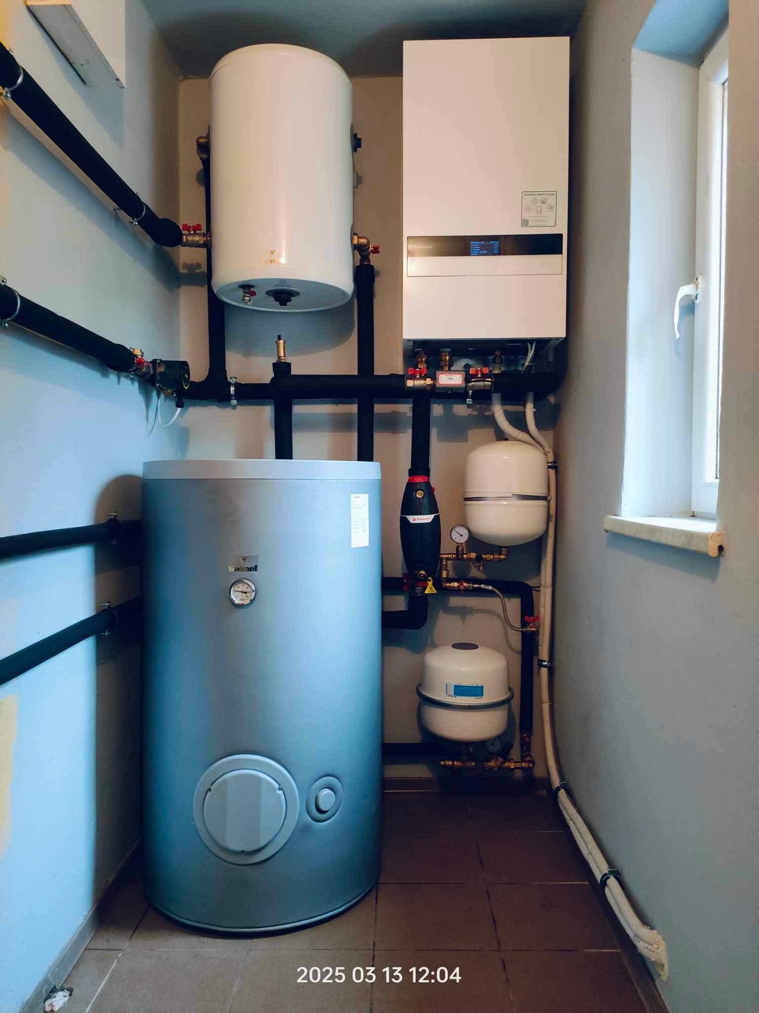 Kompletna instalacja grzewcza z piecem, zasobnikiem i pompami obiegowymi w pomieszczeniu technicznym. Widoczne rury, zawory i manometry.