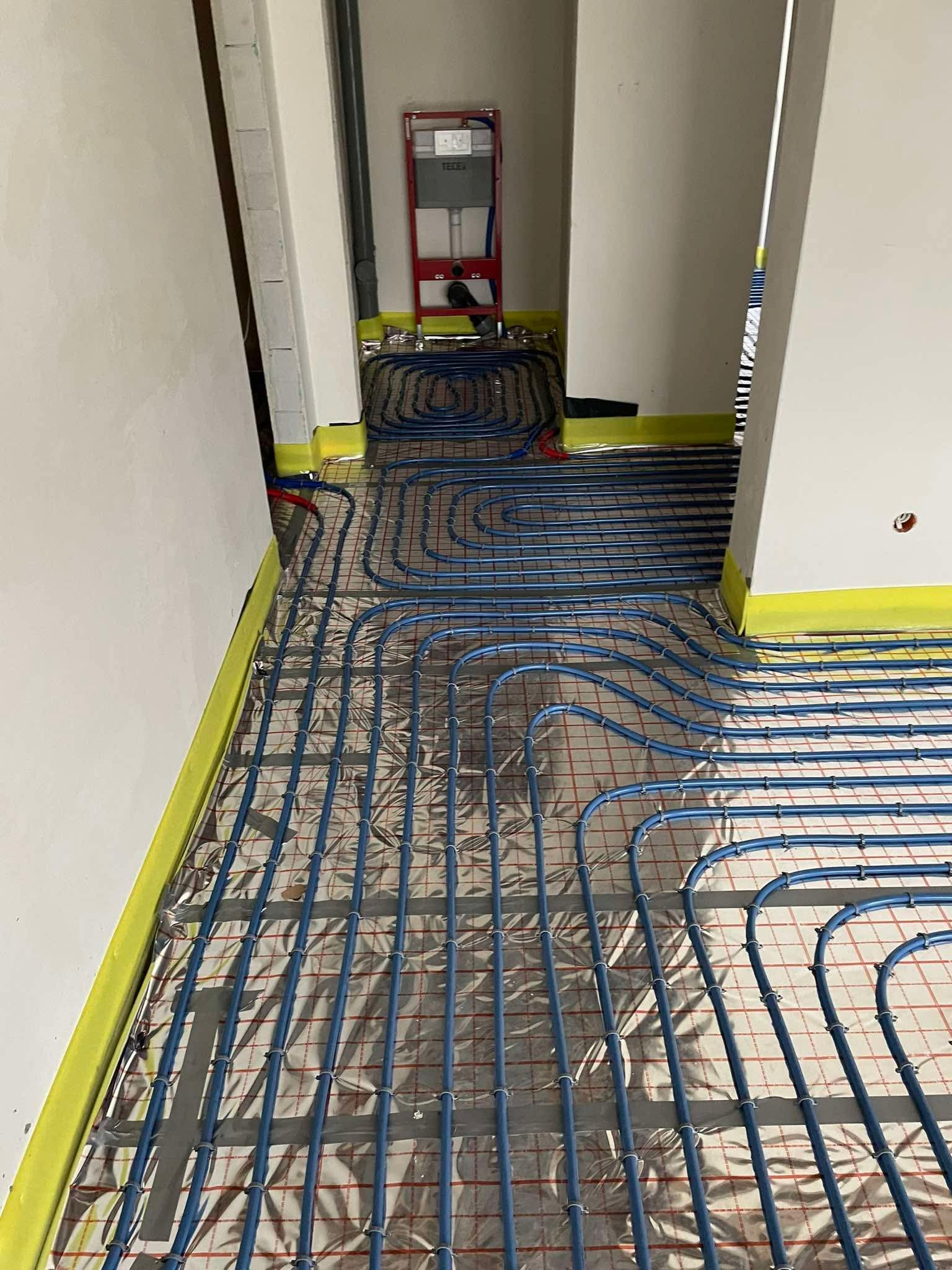 Niebieskie rury ogrzewania podłogowego ułożone na folii izolacyjnej z czerwoną siatką, przymocowane do podłoża, widok z góry, żółta taśma zabezpieczająca ściany.