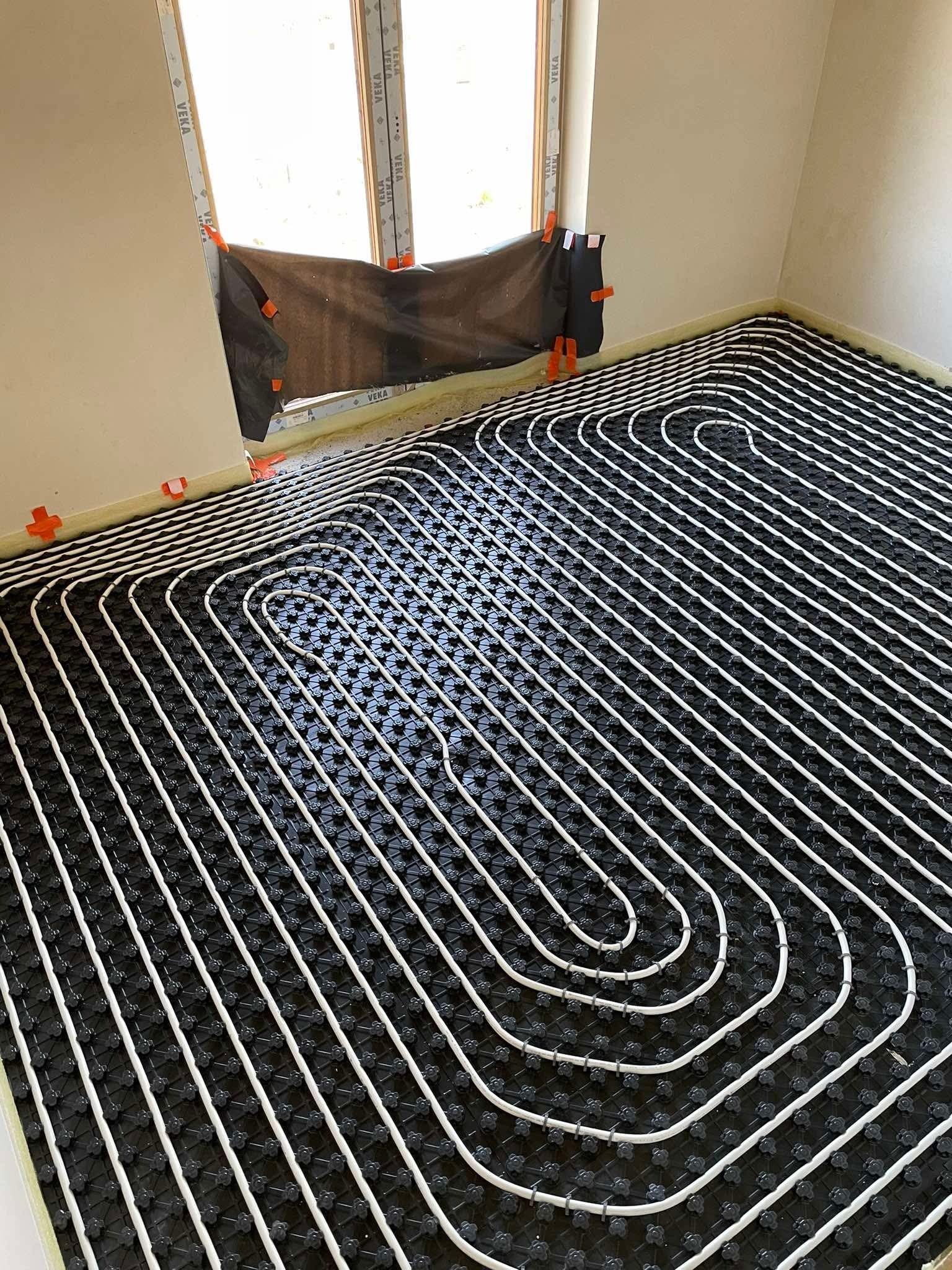 Białe rury ogrzewania podłogowego ułożone na czarnej macie w pomieszczeniu z oknem, zabezpieczonym folią. Widoczne uchwyty mocujące rury.