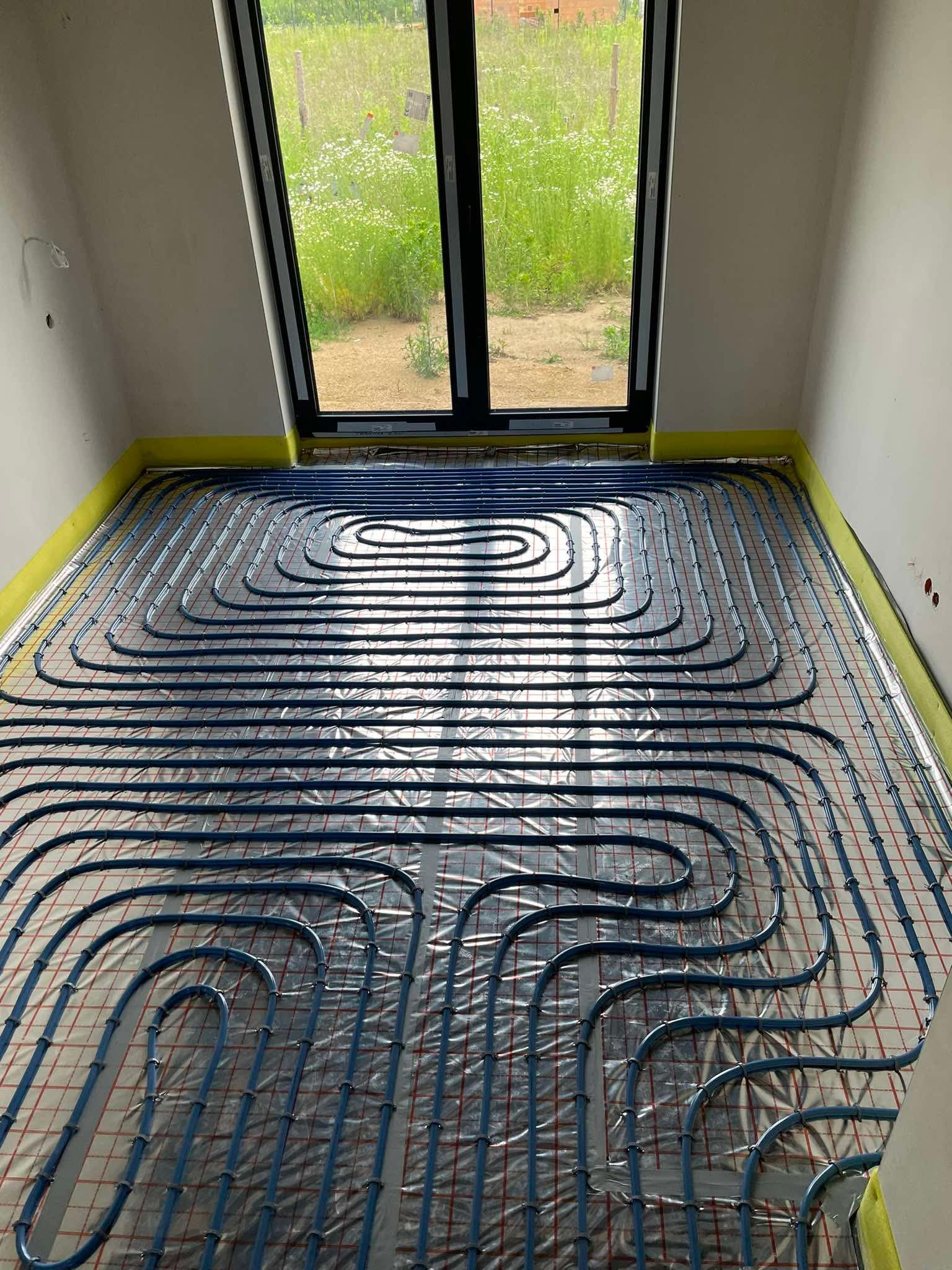 Niebieskie rury ogrzewania podłogowego ułożone równolegle na folii izolacyjnej w jasnym pokoju z oknem wychodzącym na zieleń. Żółta taśma wzdłuż ścian.