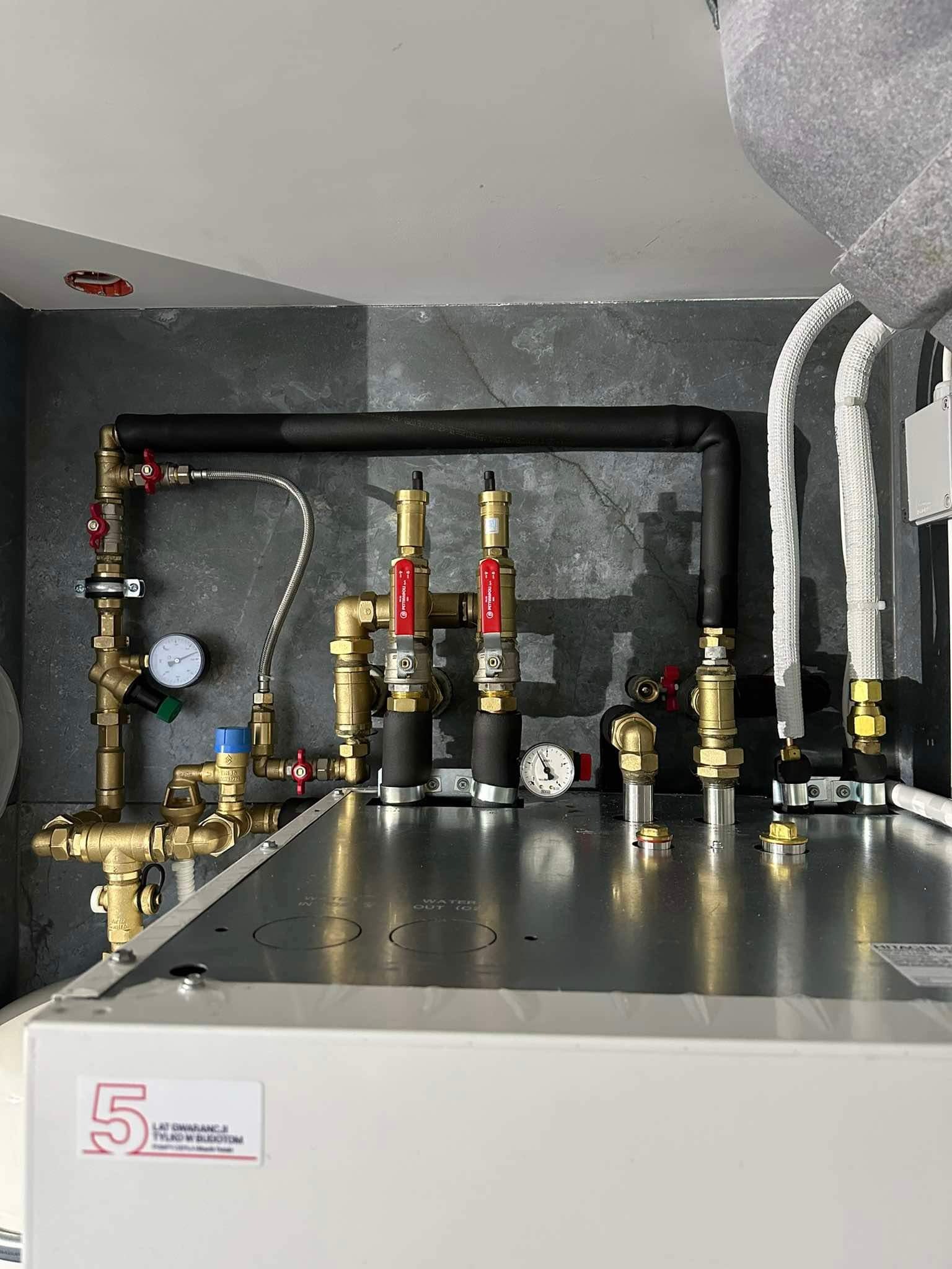 Złożona instalacja kotła grzewczego z mosiężnymi rurami, zaworami z czerwonymi uchwytami i manometrami na tle szarej ściany. Widoczna naklejka z 5-letnią gwarancją.