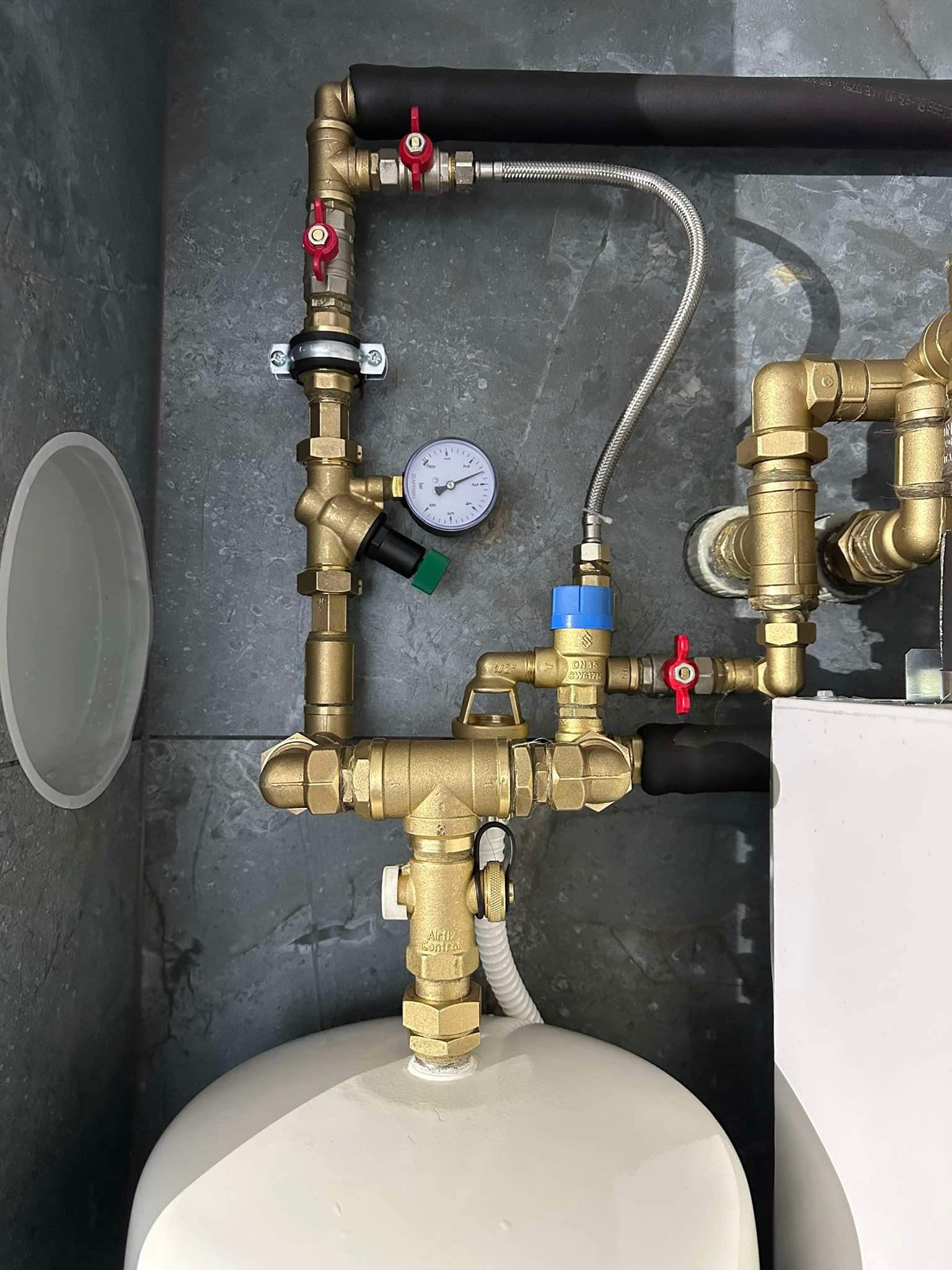 Złożona instalacja hydrauliczna z mosiężnymi złączkami, manometrem i zaworami z czerwonymi pokrętłami, podłączona do białego zbiornika w pomieszczeniu z szarą okładziną.