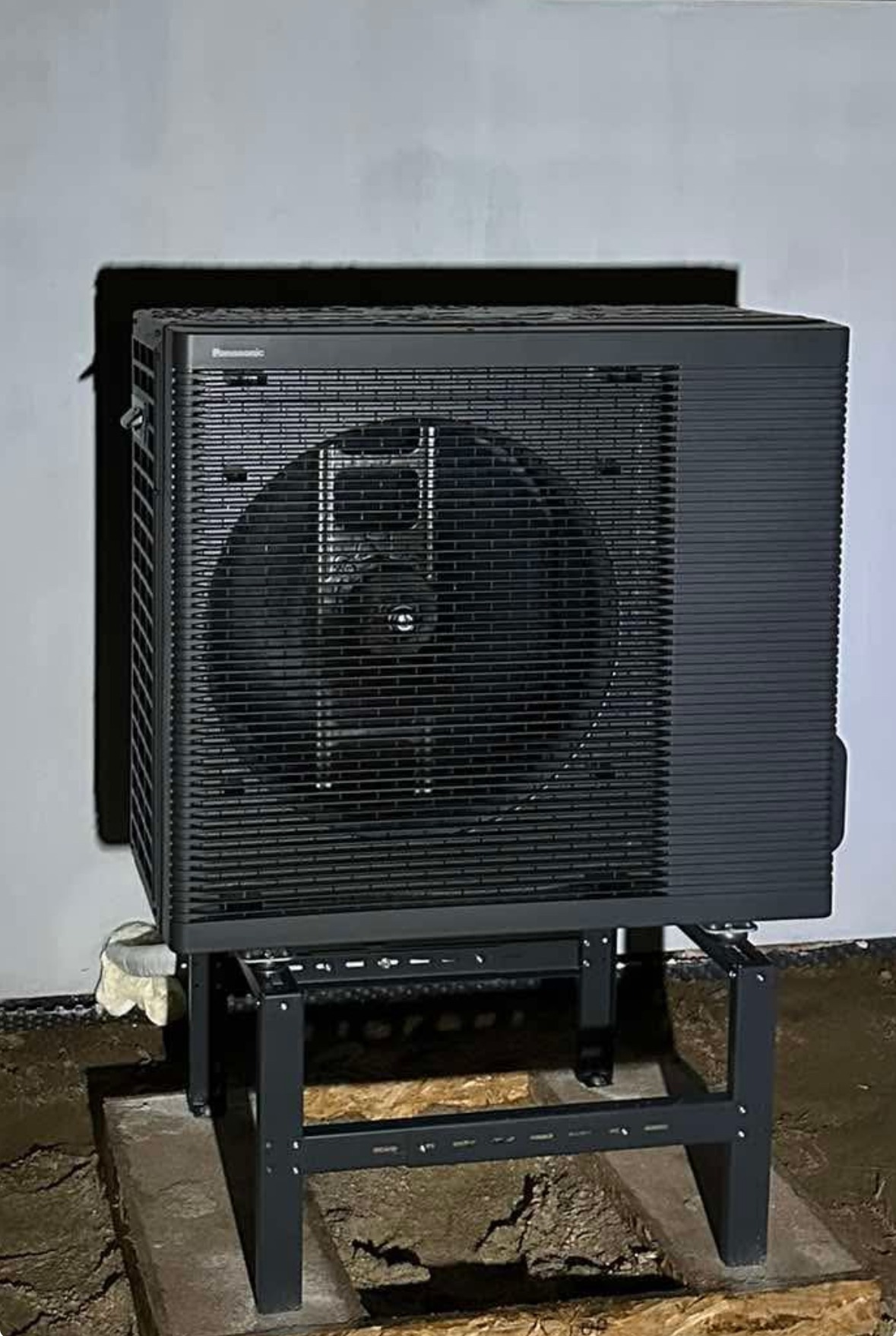 Czarna pompa ciepła Panasonic Aquarea na metalowym stelażu, zainstalowana przy białej ścianie budynku. Widoczna instalacja i ziemia wokół.