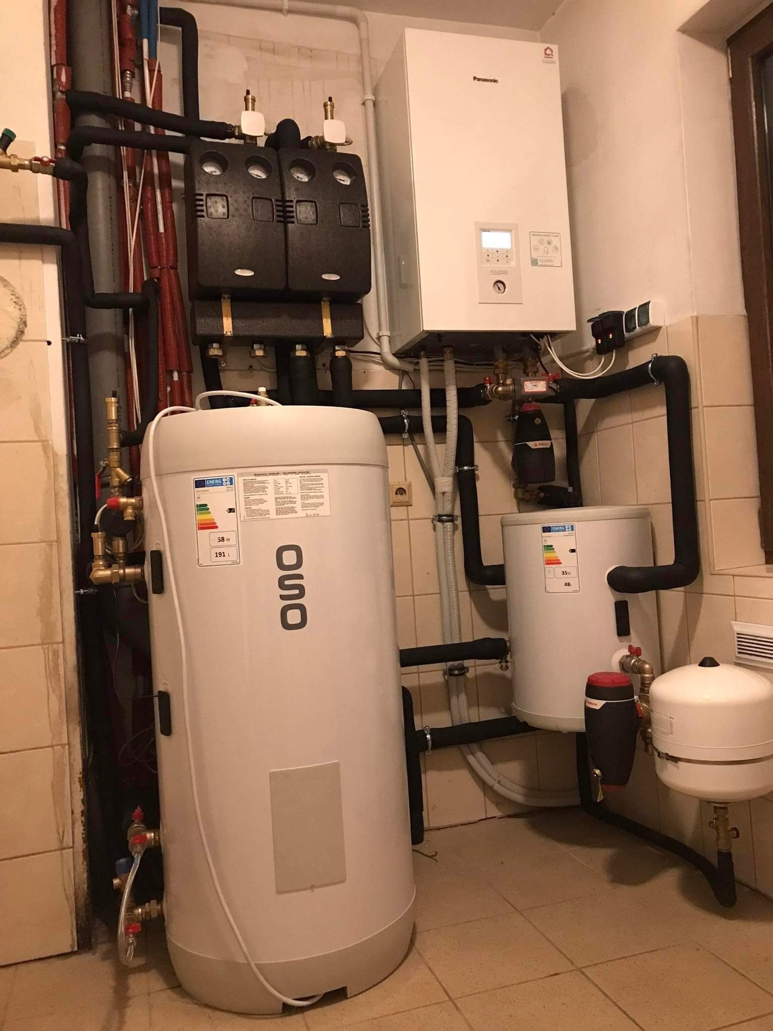 Biały bojler OSO, wiszący piec Panasonic Aquarea i czarne rury tworzą system grzewczy w pomieszczeniu z beżowymi płytkami. Widoczne zawory i manometry.