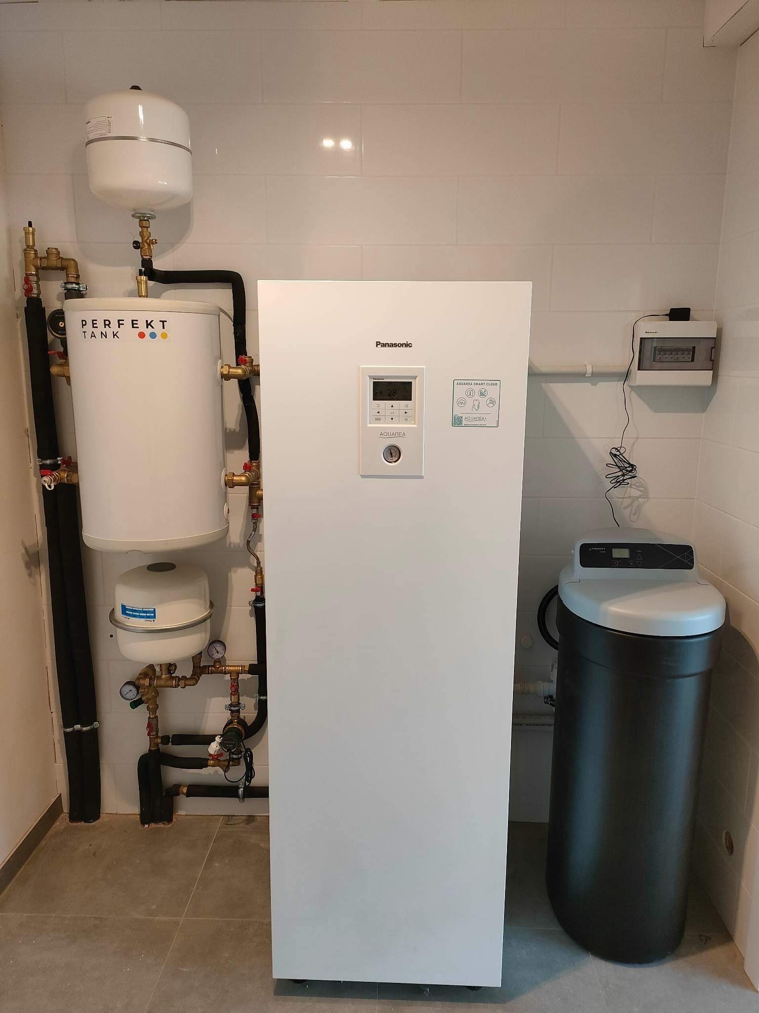 Biała pompa ciepła Panasonic Aquarea z zbiornikiem buforowym Perfekt Tank, orurowaniem i zmiękczaczem wody w pomieszczeniu z białymi płytkami.
