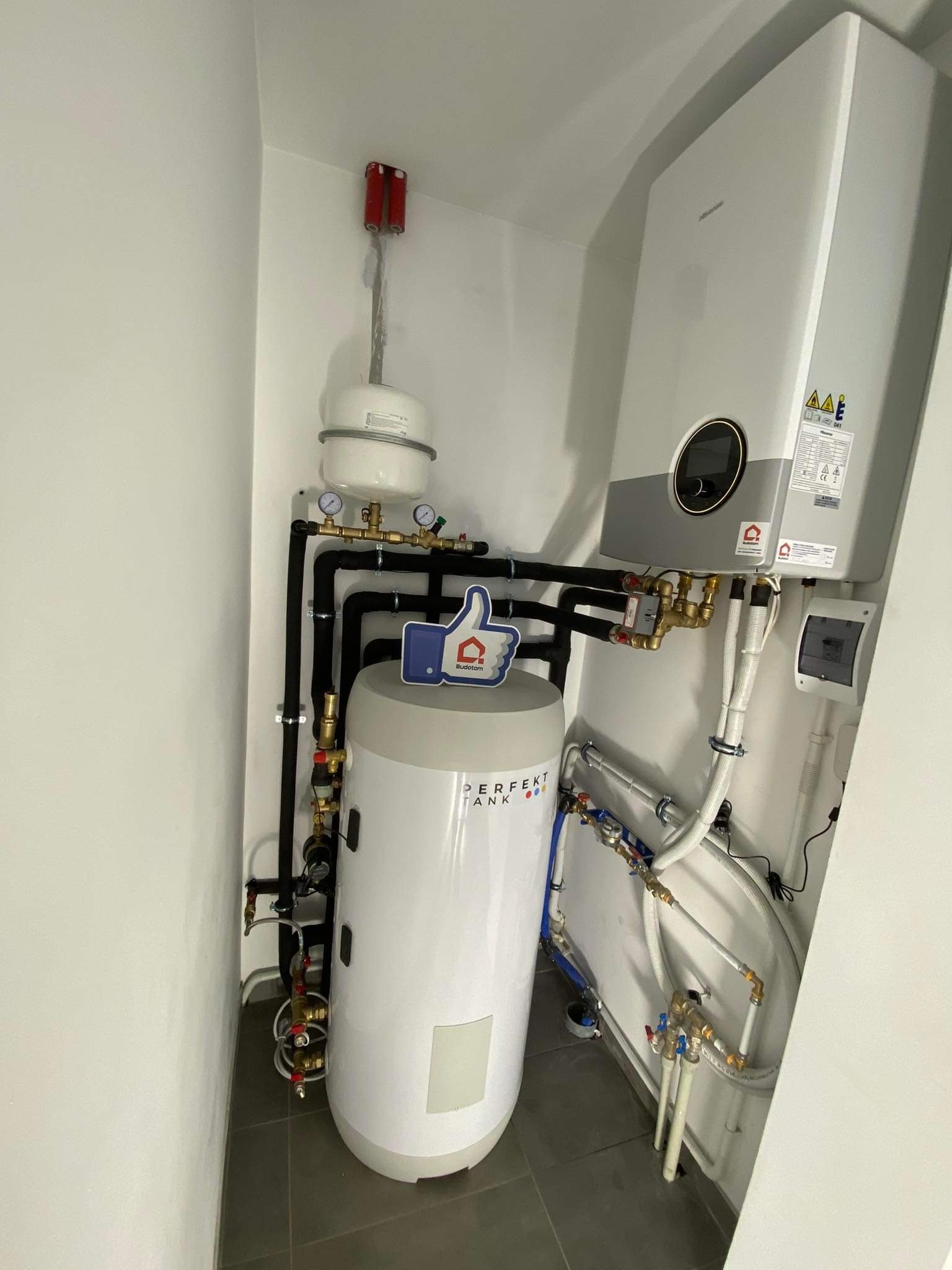 Biały zasobnik ciepłej wody użytkowej z czarnymi rurami w kotłowni, widoczne zawory, manometry i  elementy instalacji grzewczej. Na zasobniku naklejka z logotypem.