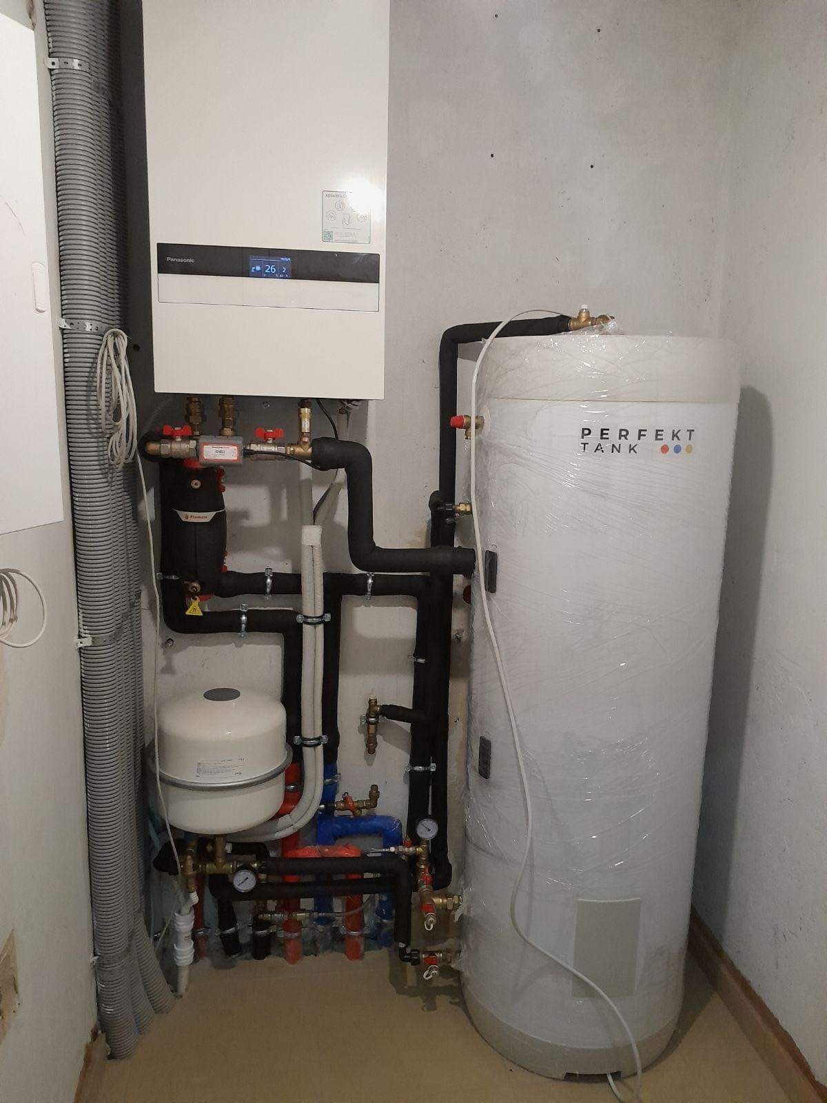 Zamontowana instalacja grzewcza z białym zasobnikiem ciepłej wody, czarnymi rurami i kotłem Panasonic. Instalacja w pomieszczeniu z szarą ścianą.