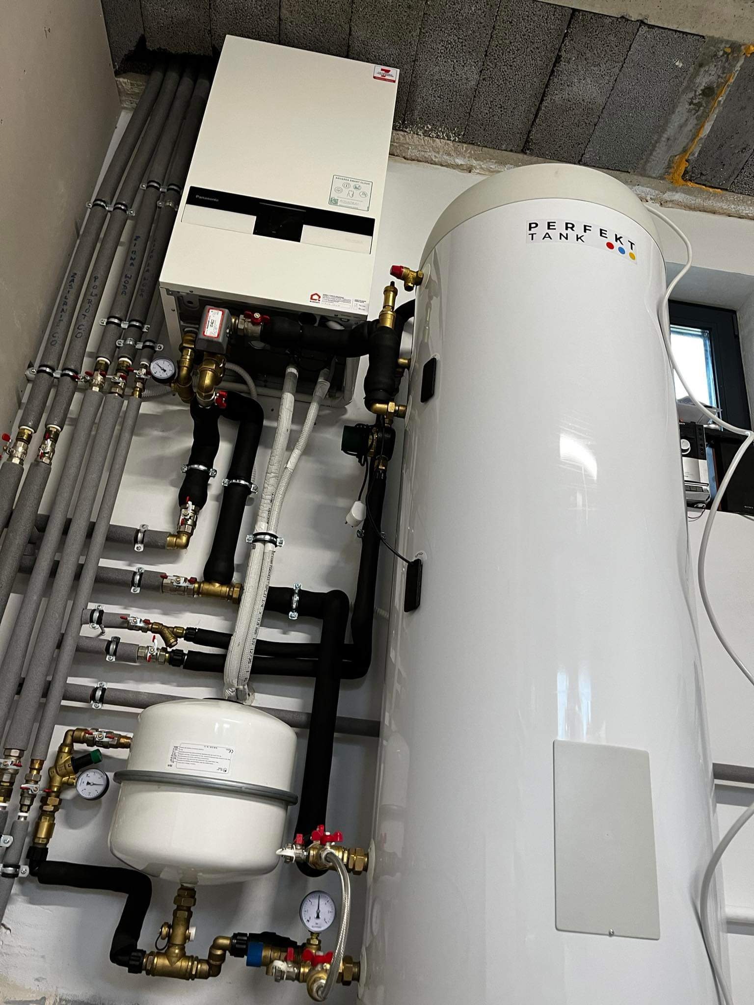 Biały zasobnik ciepłej wody użytkowej 'Perfekt Tank' i piec grzewczy z orurowaniem w kotłowni. Instalacja hydrauliczna z manometrami.