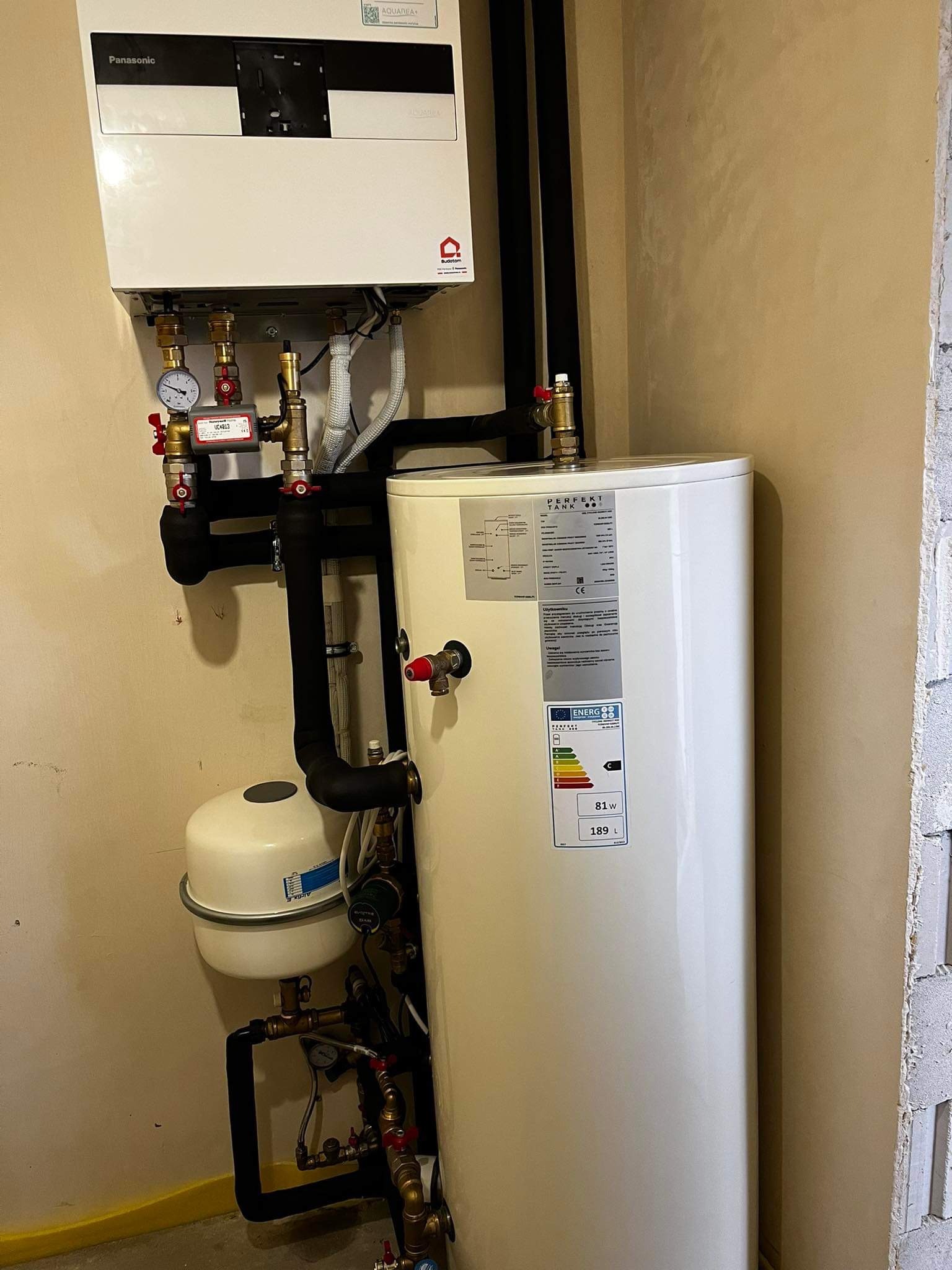 Biały, pionowy zbiornik na wodę 189L z etykietą energetyczną C, połączony czarnymi rurami z kotłem Panasonic na beżowej ścianie. Instalacja w rogu pomieszczenia.