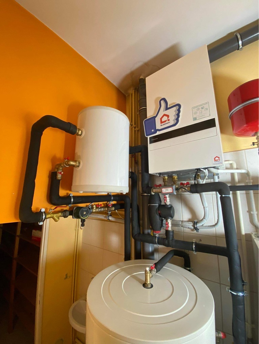 Biały zasobnik ciepłej wody i piec gazowy z czarnymi rurami w izolacji, na tle pomarańczowej ściany i beżowej glazury. Widoczne zawory i kształtki instalacyjne.