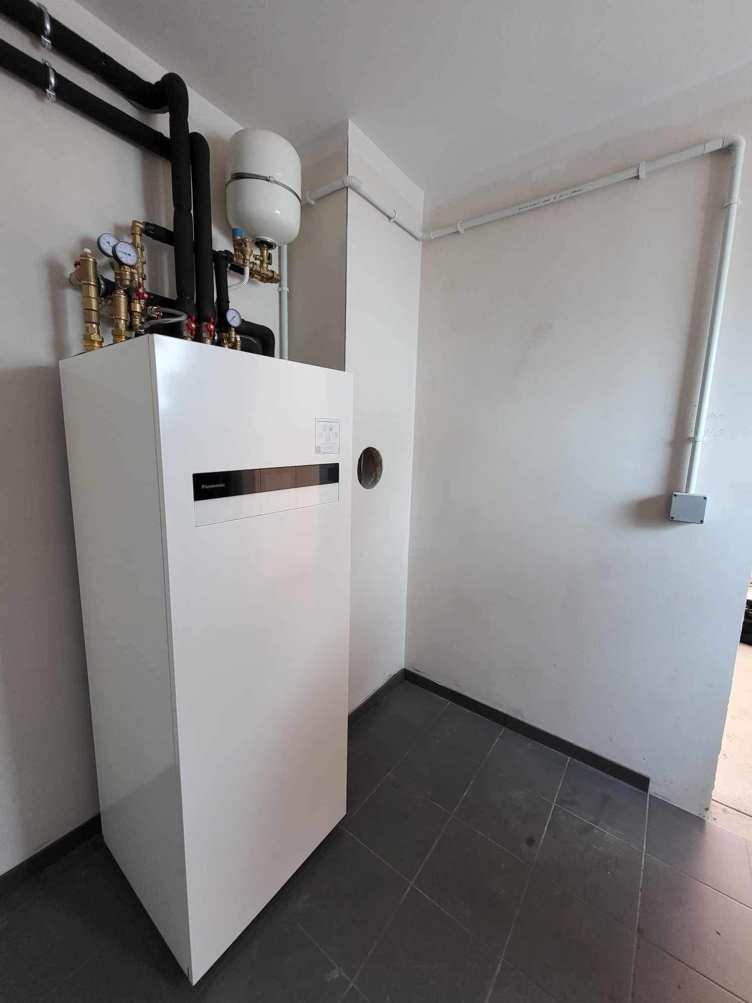 Biała pompa ciepła Panasonic z orurowaniem w pomieszczeniu technicznym we Wrocławiu. Widoczne czarne rury i manometry.