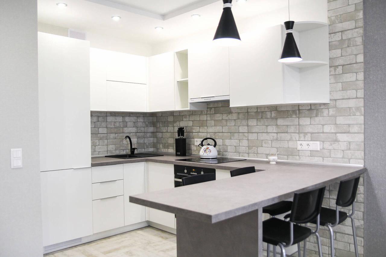 Nowoczesna kuchnia w stylu skandynawskim z białymi szafkami, szarym blatem i ceglaną ścianą, oświetlona wiszącymi lampami w kształcie stożków.