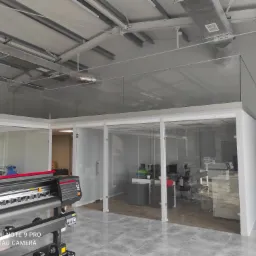 Nowoczesne biuro z przeszklonymi ścianami działowymi i widocznym ploterem wielkoformatowym, szara podłoga, surowy styl wykończenia.