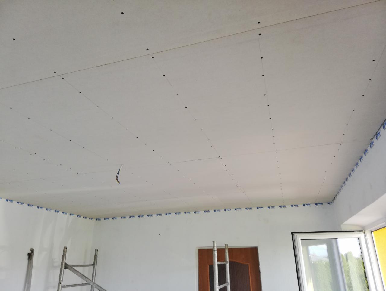 Sufit z płyt kartonowo-gipsowych w trakcie montażu, widoczne wkręty, taśma ochronna na styku ściany i sufitu, wystające przewody elektryczne.