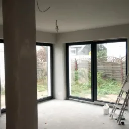 Wnętrze w trakcie prac wykończeniowych, widoczne ściany po nałożeniu warstwy wygładzającej, duże okna z czarnymi ramami wychodzące na ogród, drabina oparta o ścianę, przewody elektryczne wystające...