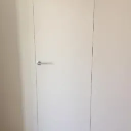 Białe drzwi ukryte w ścianie, z minimalistyczną klamką, w jasnym pomieszczeniu z drewnianą podłogą.