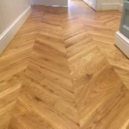 Parkiet ułożony we wzór jodełki francuskiej, wykonany z jasnego drewna dębowego, widoczny fragment ściany z białą listwą przypodłogową i szary mebel w tle.