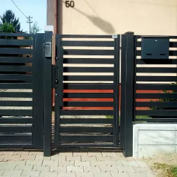 Nowoczesna, pozioma, metalowa brama wjazdowa z panelem kodowym i skrzynką na listy, w kolorze antracytowym, z elementami pomarańczowego drewna, zamontowana na betonowej podmurówce, widok z zewnątrz...