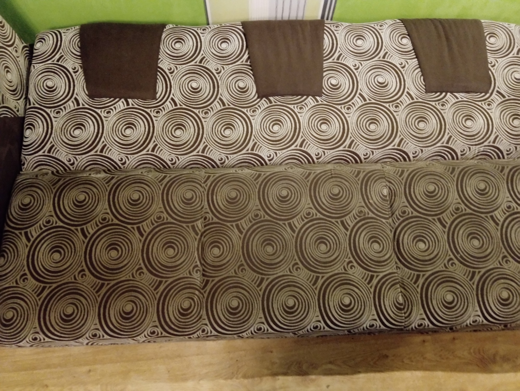 Wzór w brązowe spirale na kanapie z trzema brązowymi zagłówkami na tle zielonej ściany.