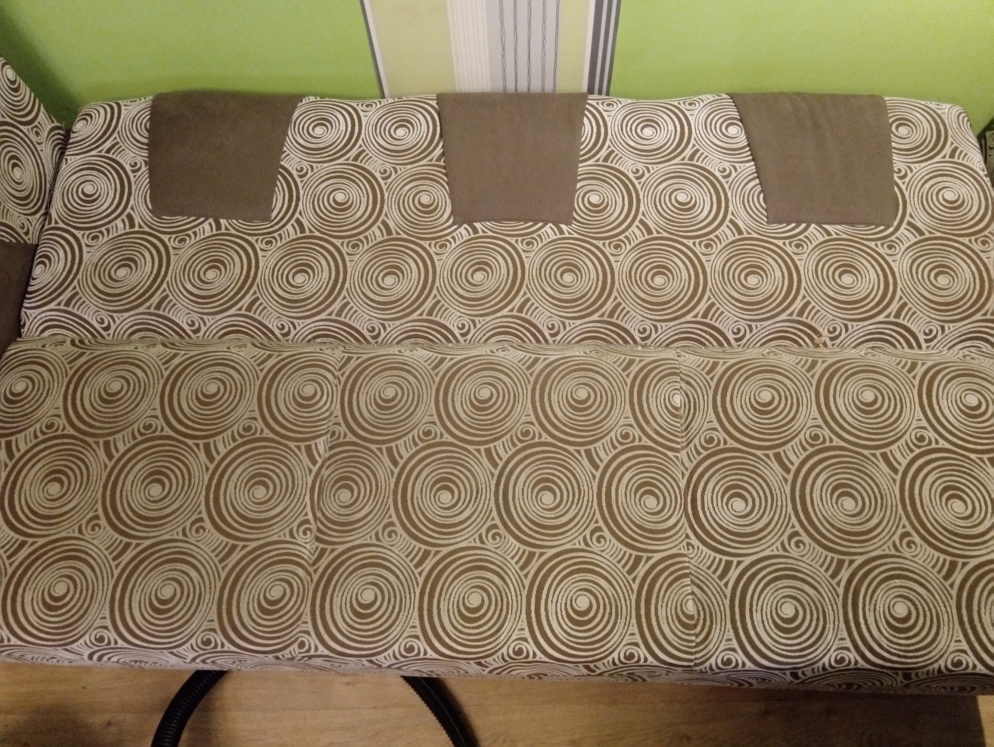 Sofa z tapicerką w brązowe i białe spirale, z trzema brązowymi zagłówkami, widoczne fragmenty podłogi i zielonej ściany.