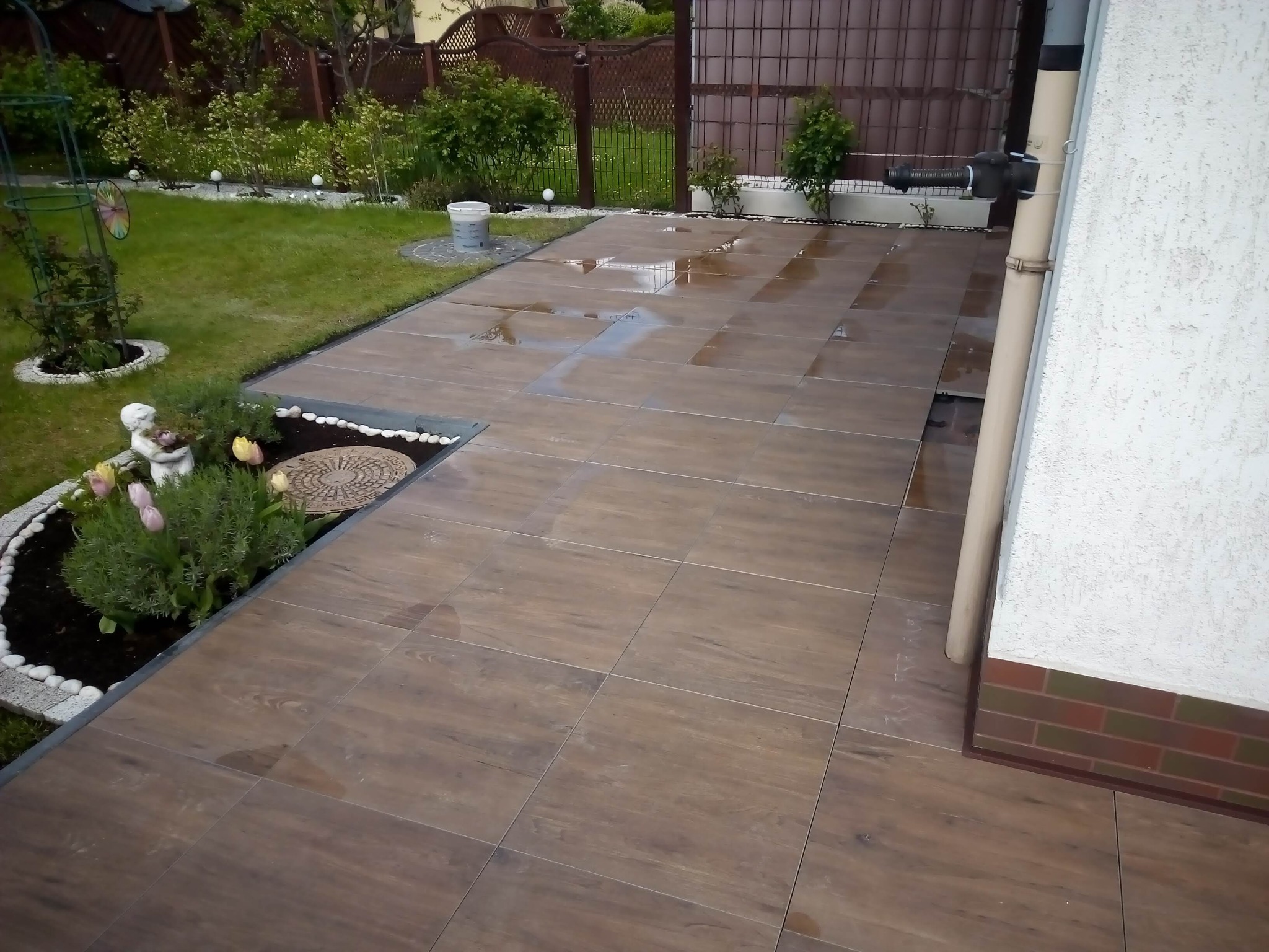 Układanie brązowych płytek imitujących drewno na tarasie przydomowym, widoczne odbicia nieba na mokrej powierzchni, obok rabata kwiatowa z białymi kamieniami i dekoracyjną figurką.