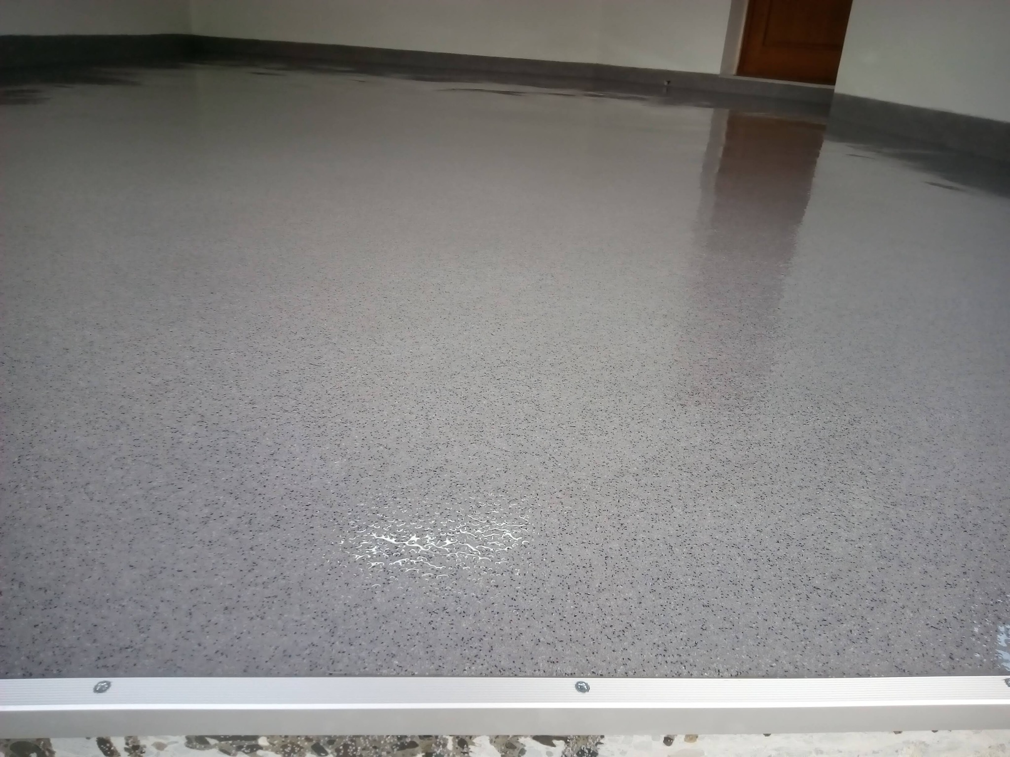 Świeżo wylana, szara posadzka z drobnymi, ciemnymi plamkami, odbijająca światło, z widoczną listwą przypodłogową i fragmentem drzwi w tle.