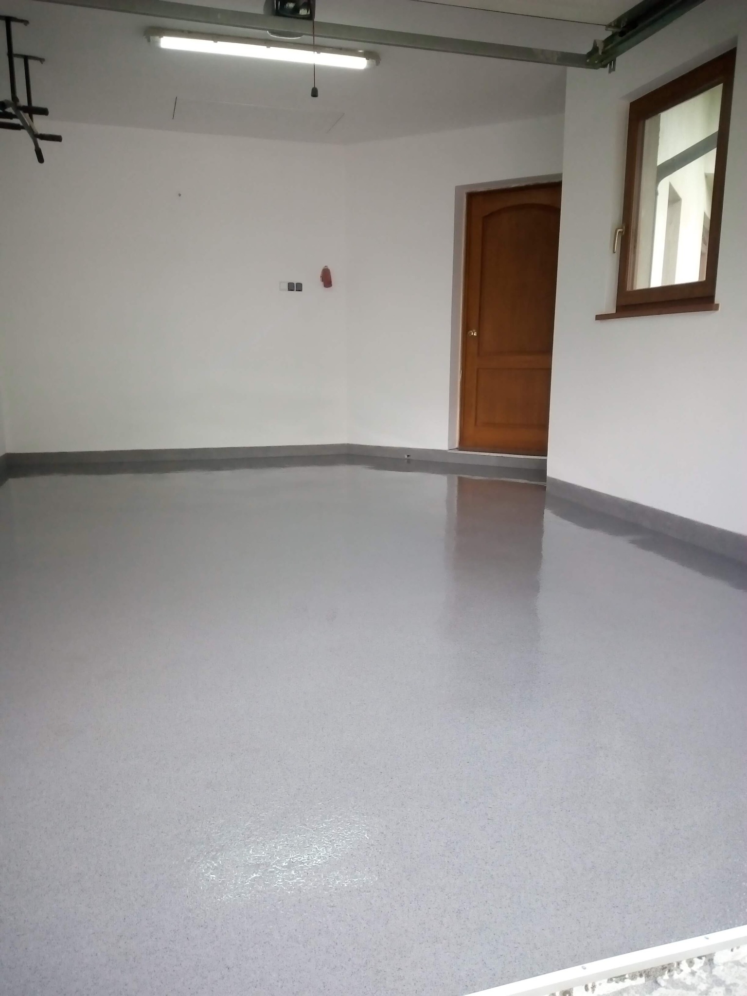 Szara, błyszcząca posadzka z żywicy epoksydowej w garażu w Żywcu, widoczna drewniana brama i okno, białe ściany, surowy wystrój.