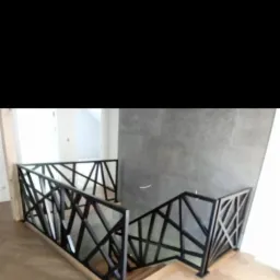 Nowoczesna, czarna balustrada schodowa o geometrycznym wzorze na drewnianych schodach, z szarą ścianą w tle.