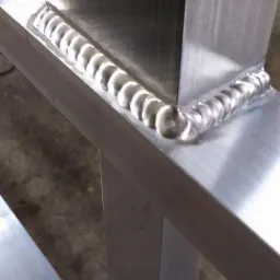 Szczegółowe zbliżenie na świeżo wykonane spawy łączące elementy konstrukcji ze stali nierdzewnej; widoczne regularne, srebrzyste ślady spawania wzdłuż krawędzi.