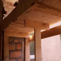 Wnętrze w trakcie prac budowlanych: drewniana konstrukcja podtrzymująca strop, surowa cegła, widoczne elementy instalacji.