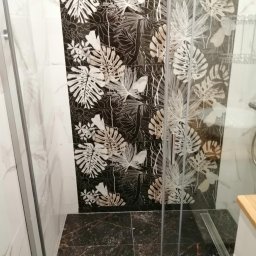 Przedsiębiorstwo usługowo handlowe - Kabina prysznicowa z czarnymi płytkami w kwiatowe wzory, marmurowa podłoga i białe ściany z efektem marmuru.