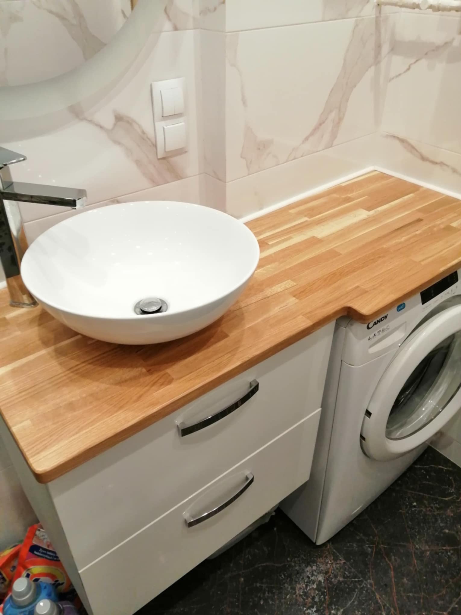 Nowoczesna łazienka z umywalką nablatową, drewnianym blatem nad pralką Candy i marmurowymi płytkami na ścianie.